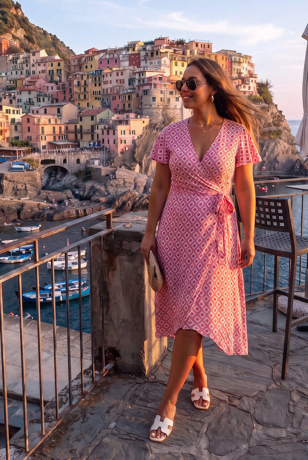 Una mujer que luce el elegante vestido largo de verano cruzado de Lohe, con estampado de rombos y gafas de sol, posa en una terraza con vistas panorámicas a los emblemáticos edificios de colores situados en los acantilados y a los barcos en el mar al atardecer, ideal para resaltar el estilo y la moda veraniega en entornos costeros.