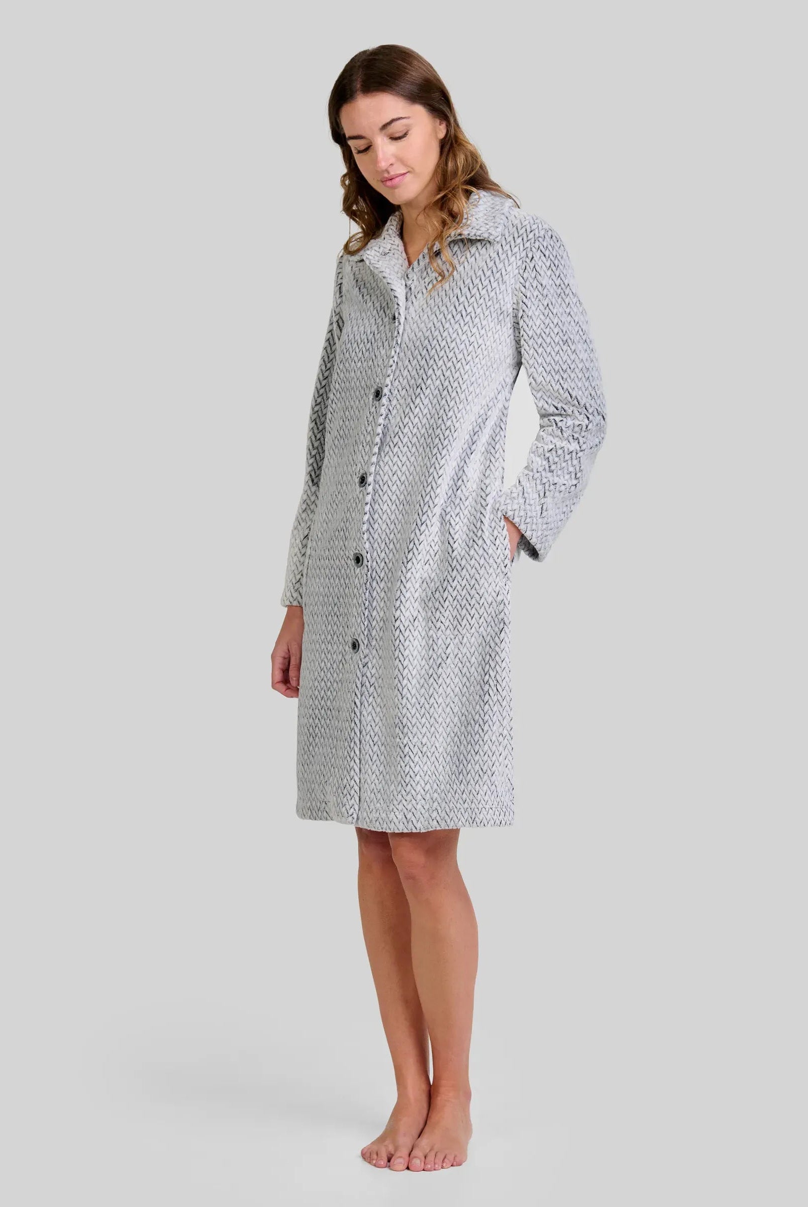 Mujer de pie descalza con bata larga gris claro y textura espiga, botones al frente y manos en los bolsillos