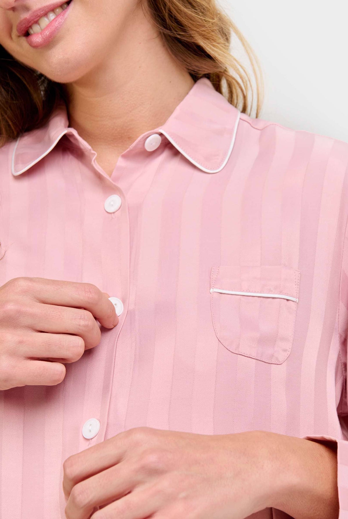 Mujer abrochándose chaquetilla abotonada de pijama rosa palo de rayas, ribete y botones blancos