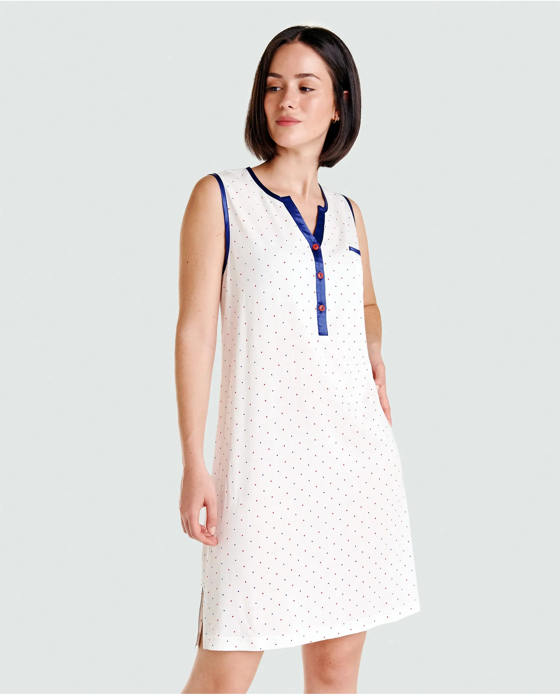 A woman wears an elegant, sleeveless, knee-length, white nightdress, decorated with delicate dots and elegant dark blue piping.