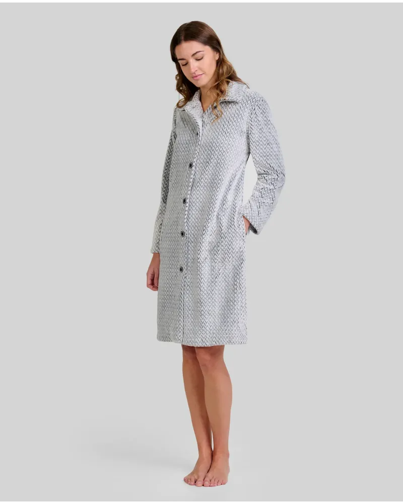 Woman standing barefoot wearing a long light grey herringbone robe with buttons down the front and her hands in her pockets.