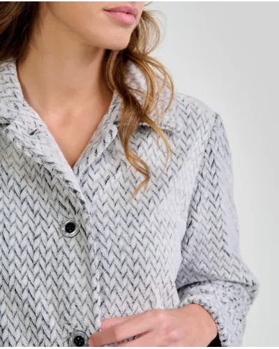 Woman standing barefoot wearing a long light grey herringbone robe with buttons down the front and her hands in her pockets.