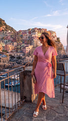 Una mujer que luce el elegante vestido largo de verano cruzado de Lohe, con estampado de rombos y gafas de sol, posa en una terraza con vistas panorámicas a los emblemáticos edificios de colores situados en los acantilados y a los barcos en el mar al atardecer, ideal para resaltar el estilo y la moda veraniega en entornos costeros.