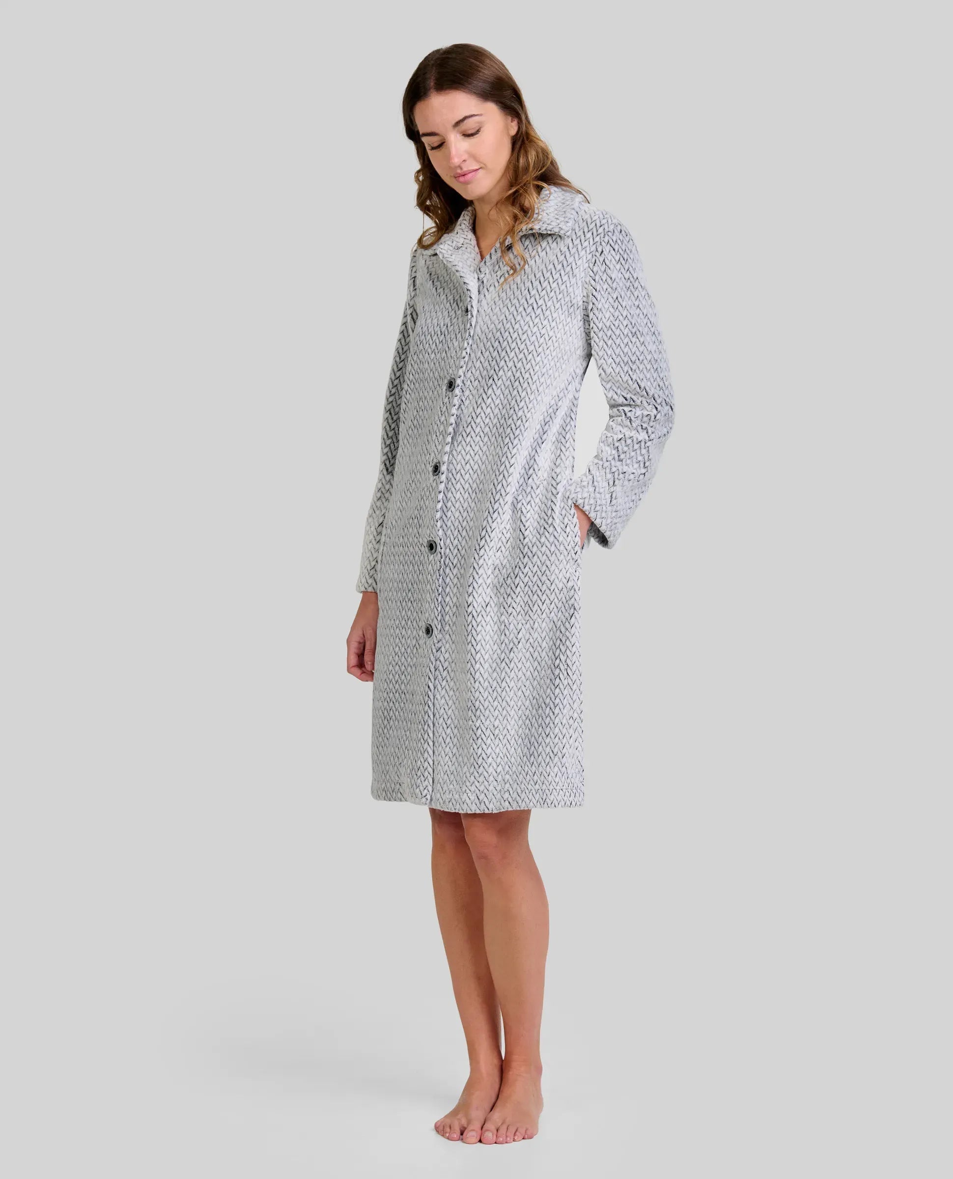 Mujer de pie descalza con bata larga gris claro y textura espiga, botones al frente y manos en los bolsillos