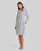 Mujer de pie descalza con bata larga gris claro y textura espiga, botones al frente y manos en los bolsillos