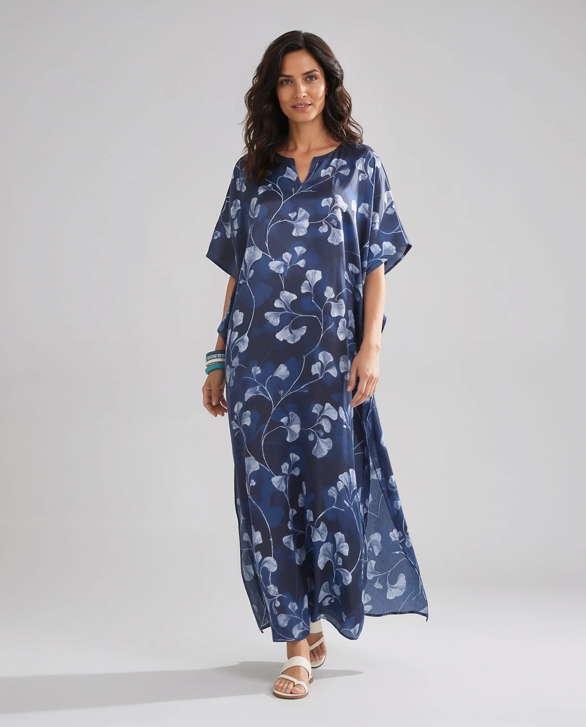 Mujer de pie con un kaftan de seda natural largo azul de estampado floral y sandalias blancas.