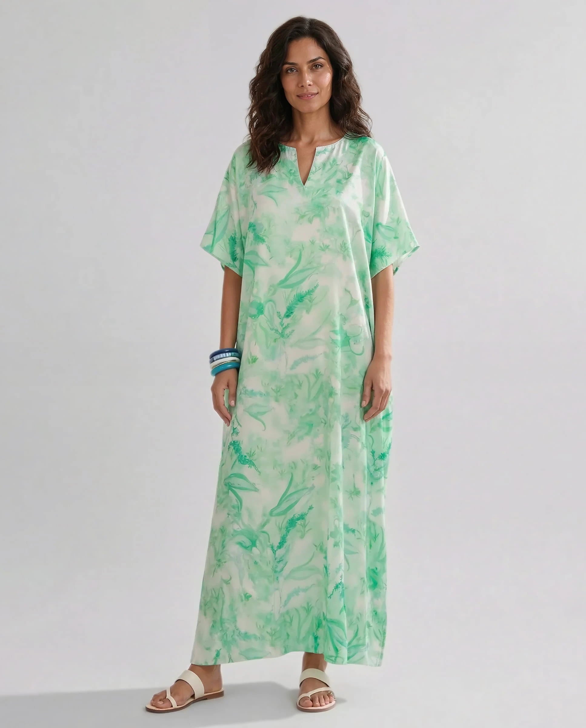 Mujer de pie con caftan largo hasta los tobillos de estampado verde y sandalias blancas sobre fondo claro.