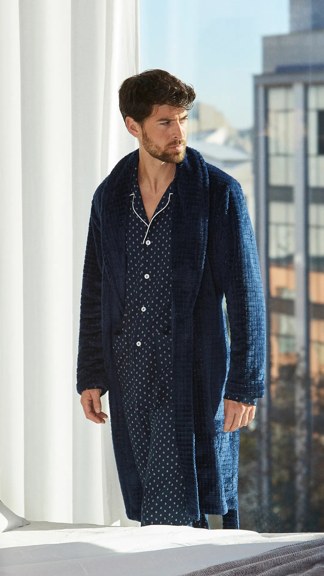 Hombre con bata azul marino y pijama de abierto de pequeños cachemires, de pie junto a ventana