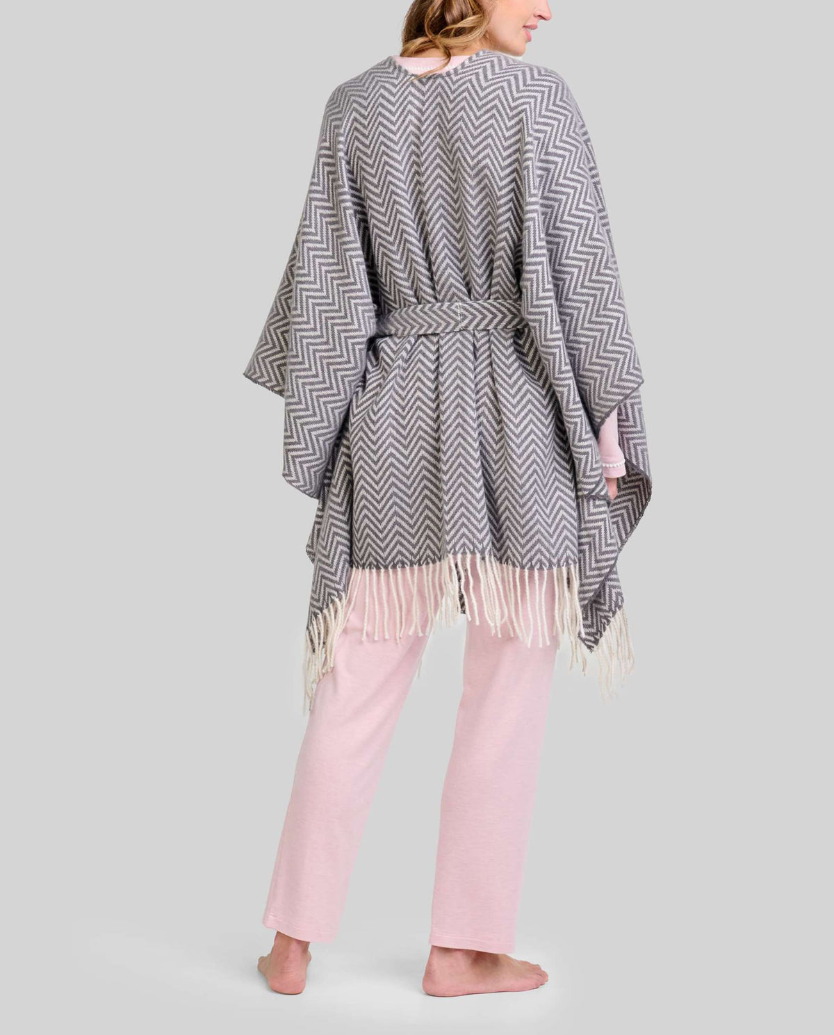 Mujer de espaldas con poncho gris de zigzag y flecos, sobre pijama rosa de invierno